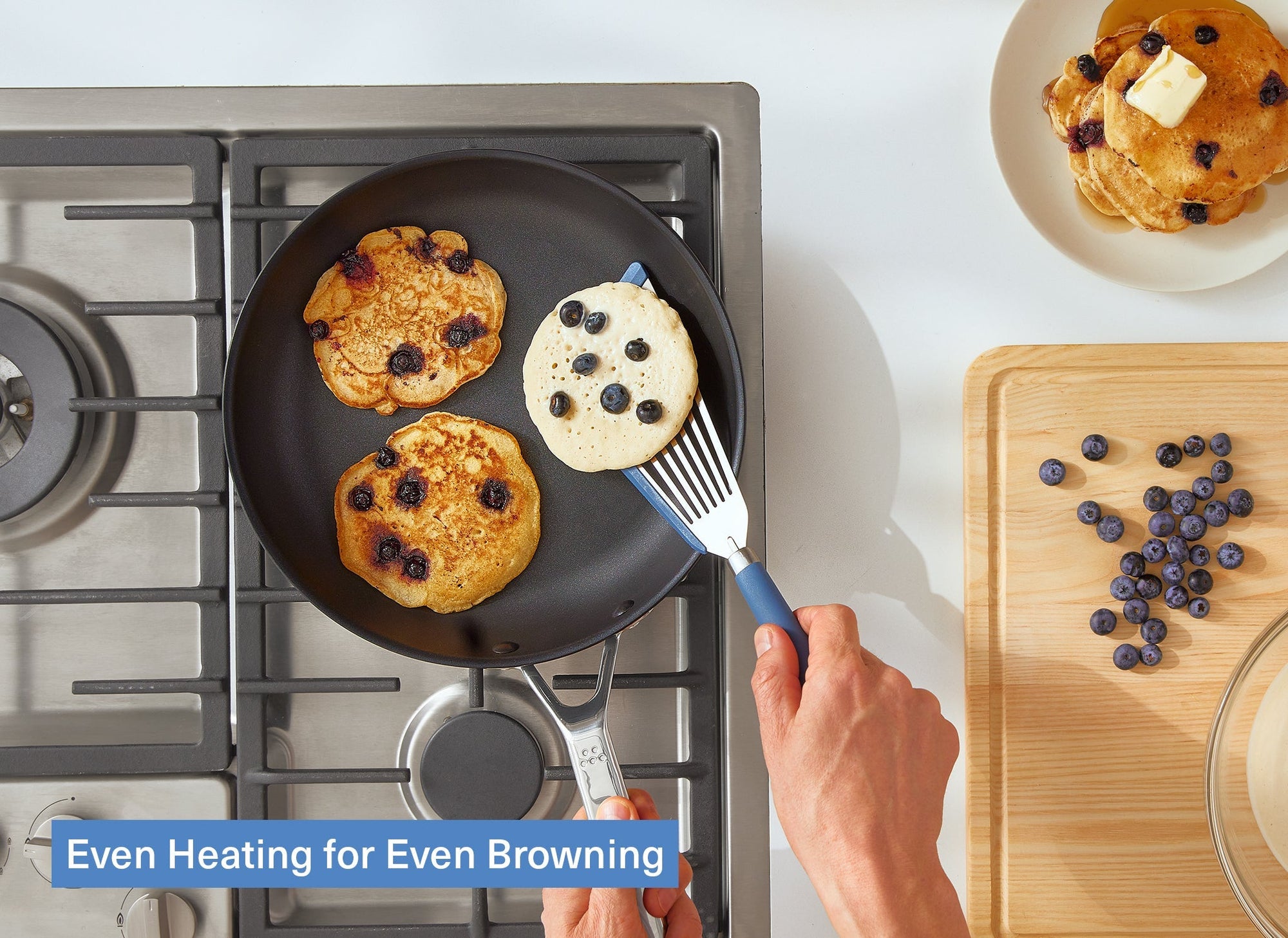 A bird’s eye view of a Misen Nonstick Pan on a stovetop with a chef’s hand flipping blueberry pancakes in it. A stack of pancakes sits on a plate to the right of the stovetop. A caption reads: “Even Heating for Even Browning.”