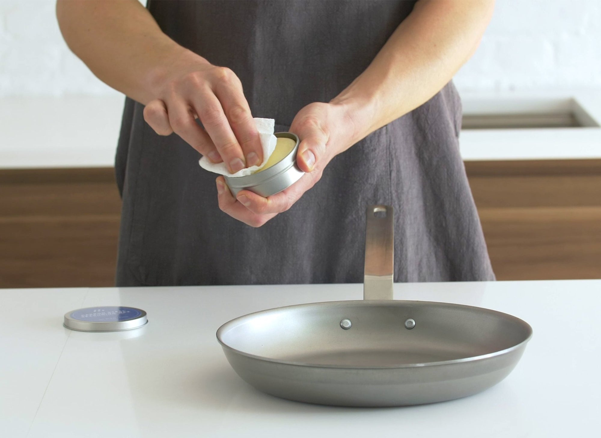 {{3-25-oz}} Hands wiping seasoning wax puck with a paper towel held above an unseasoned carbon steel pan on a countertop.