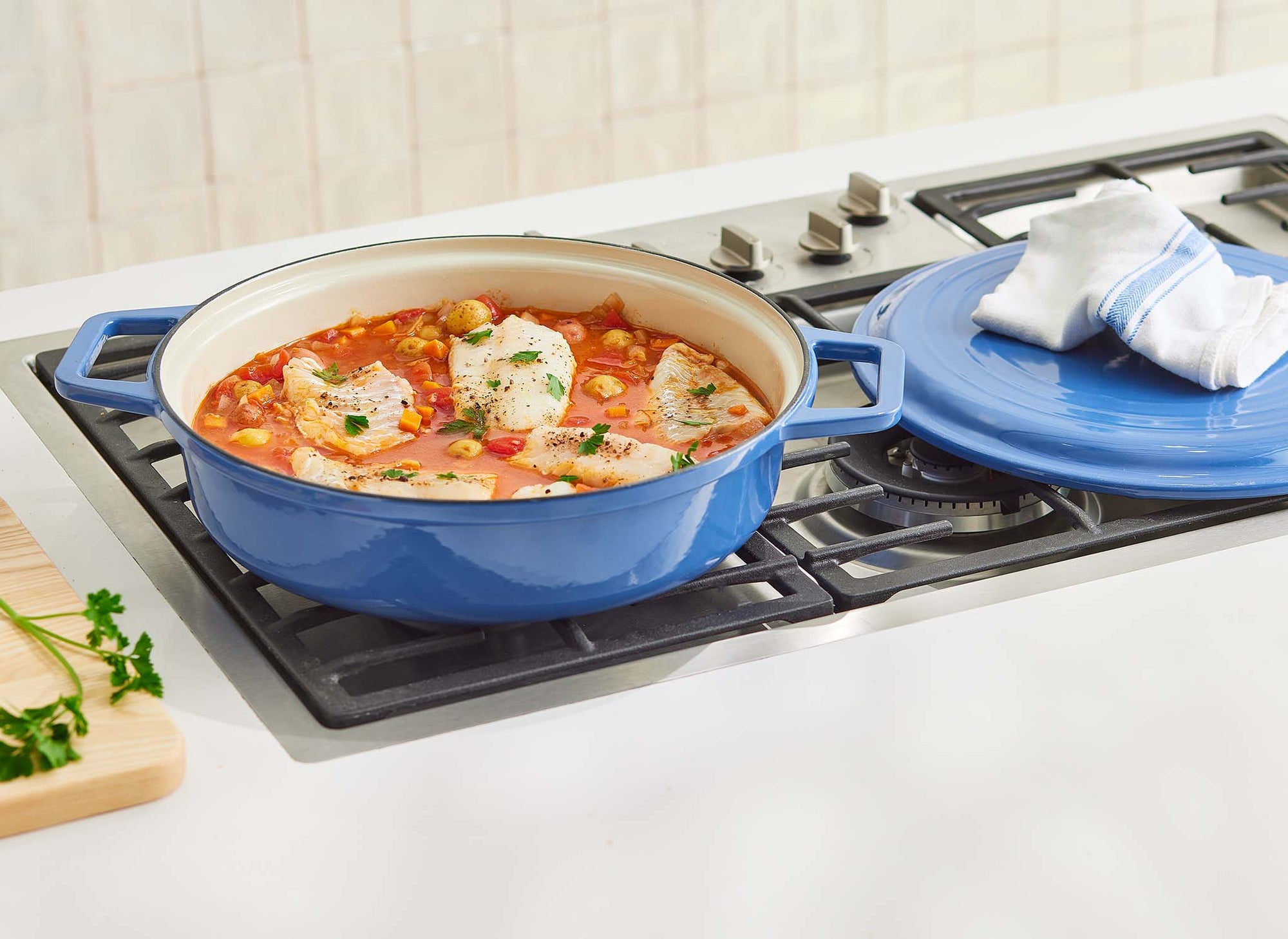 {{blue, black, gray, red, green}} A blue Misen Enameled Cast Iron Braiser with lid removed, revealing a braised fish and tomato sauce dish, ready to be cooked.