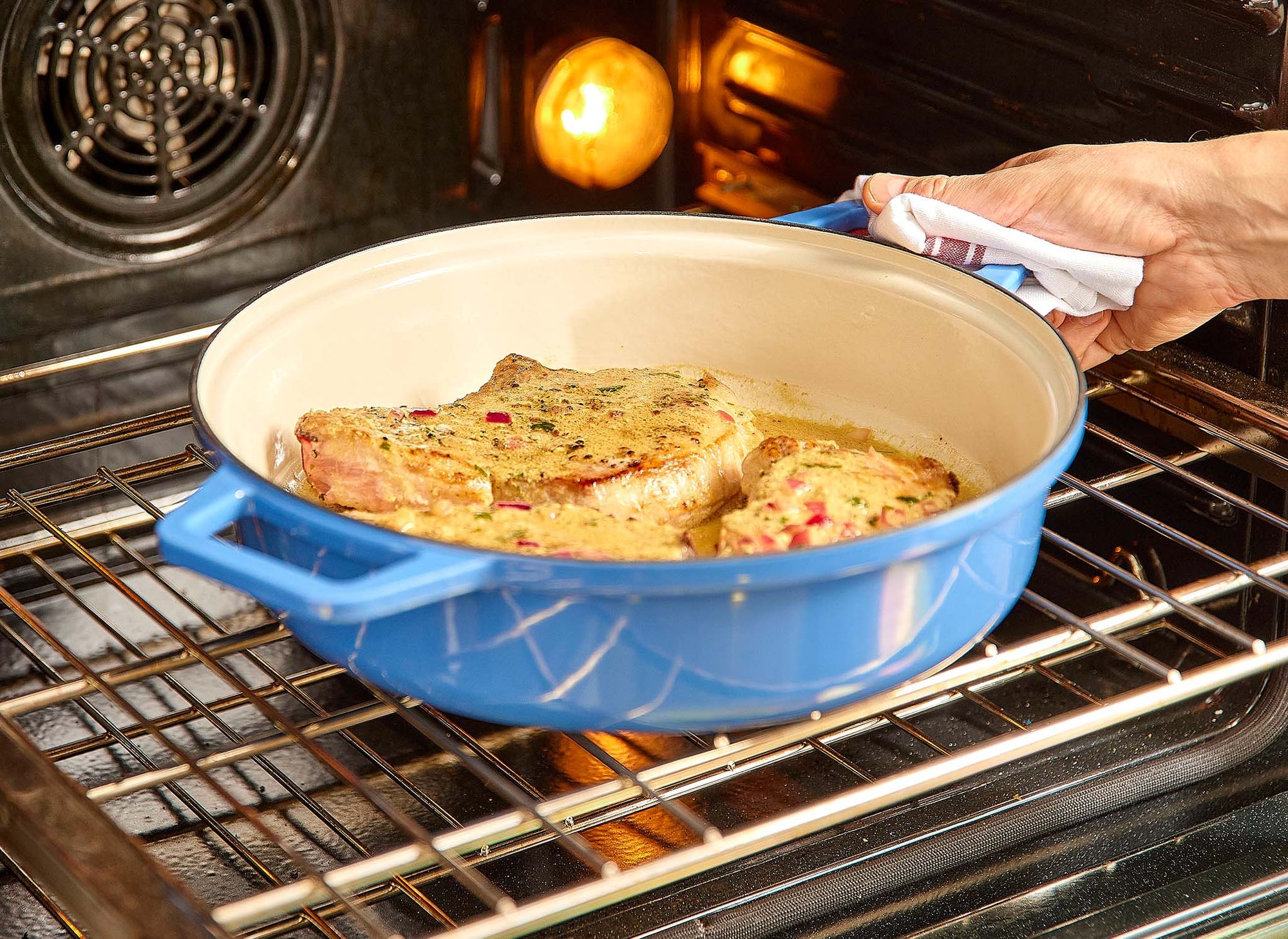 {{blue, black, gray, red, green}} A chef places a blue Misen Enameled Cast Iron Braiser containing seared pork chops into an oven to finish cooking