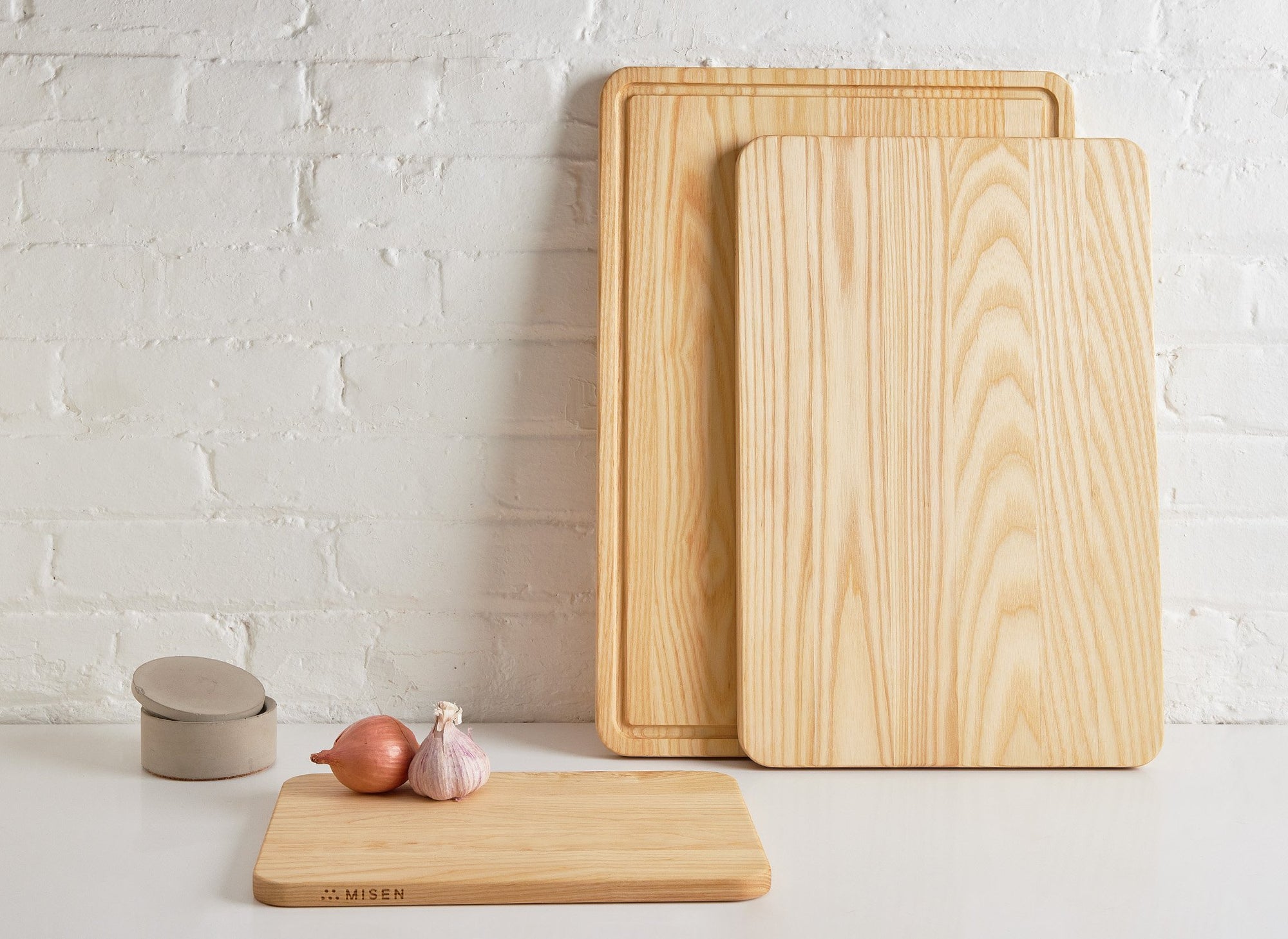 {{small,medium,large}} A Small Misen Wood Cutting Board flat on a countertop, with Medium and Large Wood Cutting Boards leaning against a white brick wall.