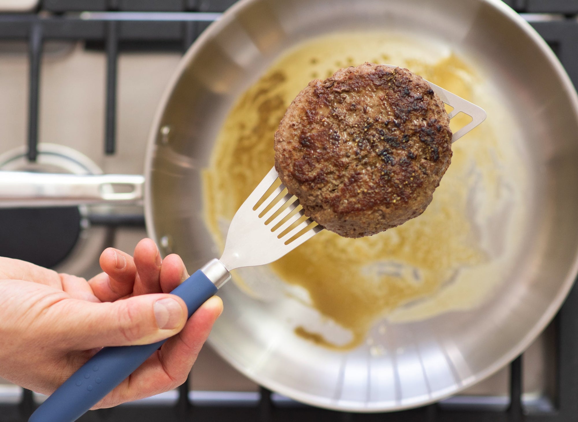 {{stainless-steel-blue,stainless-steel-black,stainless-steel-gray}} A Misen Metal Fish Spatula with blue silicone handle lifting a cooked hamburger patty above a stainless steel skillet.