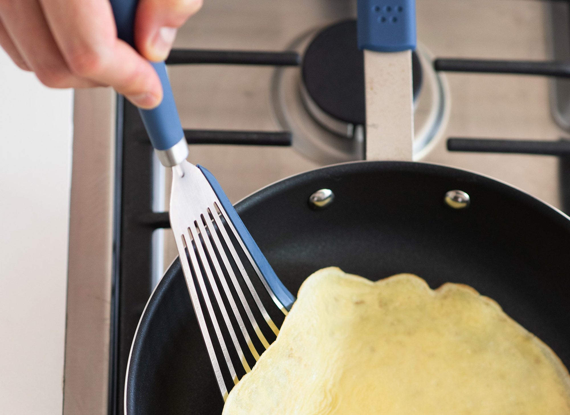 {{silicone-blue,silicone-black,silicone-gray}} A Blue Misen Silicone Fish Spatula gently folding a thin french omelet inside a nonstick pan on a stovetop.