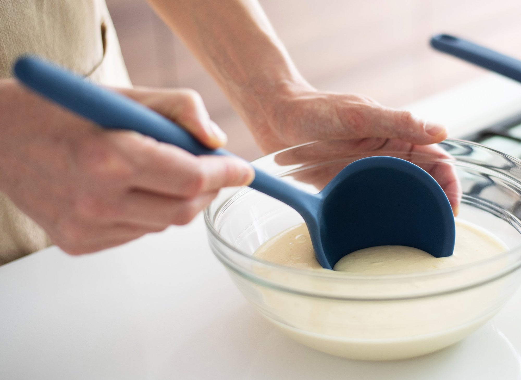 {{blue,black,gray}} Hand using Blue Misen Ladle to scoop batter from a glass mixing bowl on a white kitchen countertop.