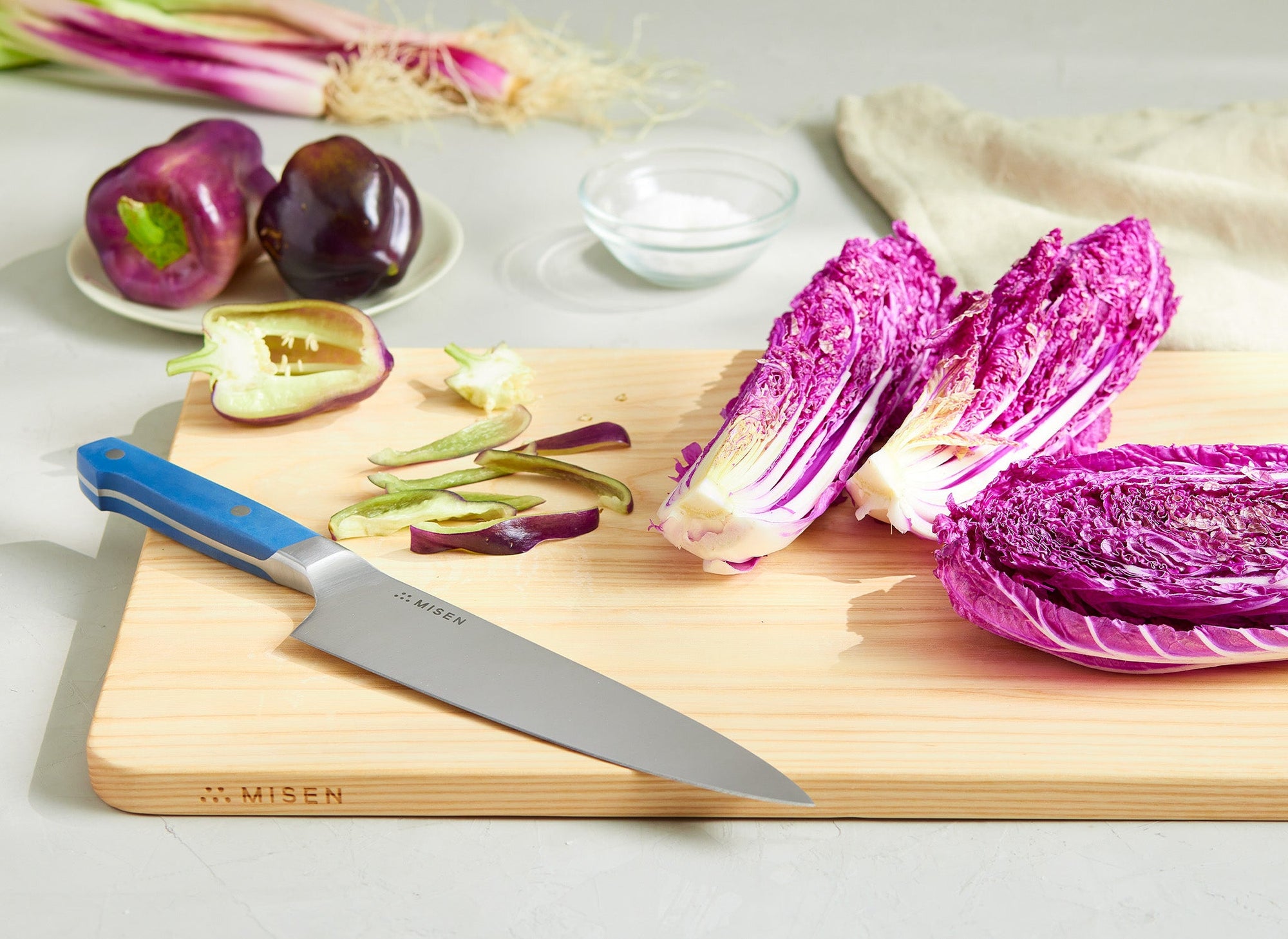 {{blue}} A Misen Chef’s Knife with a blue handle rests at an angle on a wood Misen cutting board. Next to the knife are assorted chopped vegetables such as lettuce, bell peppers, and onions.