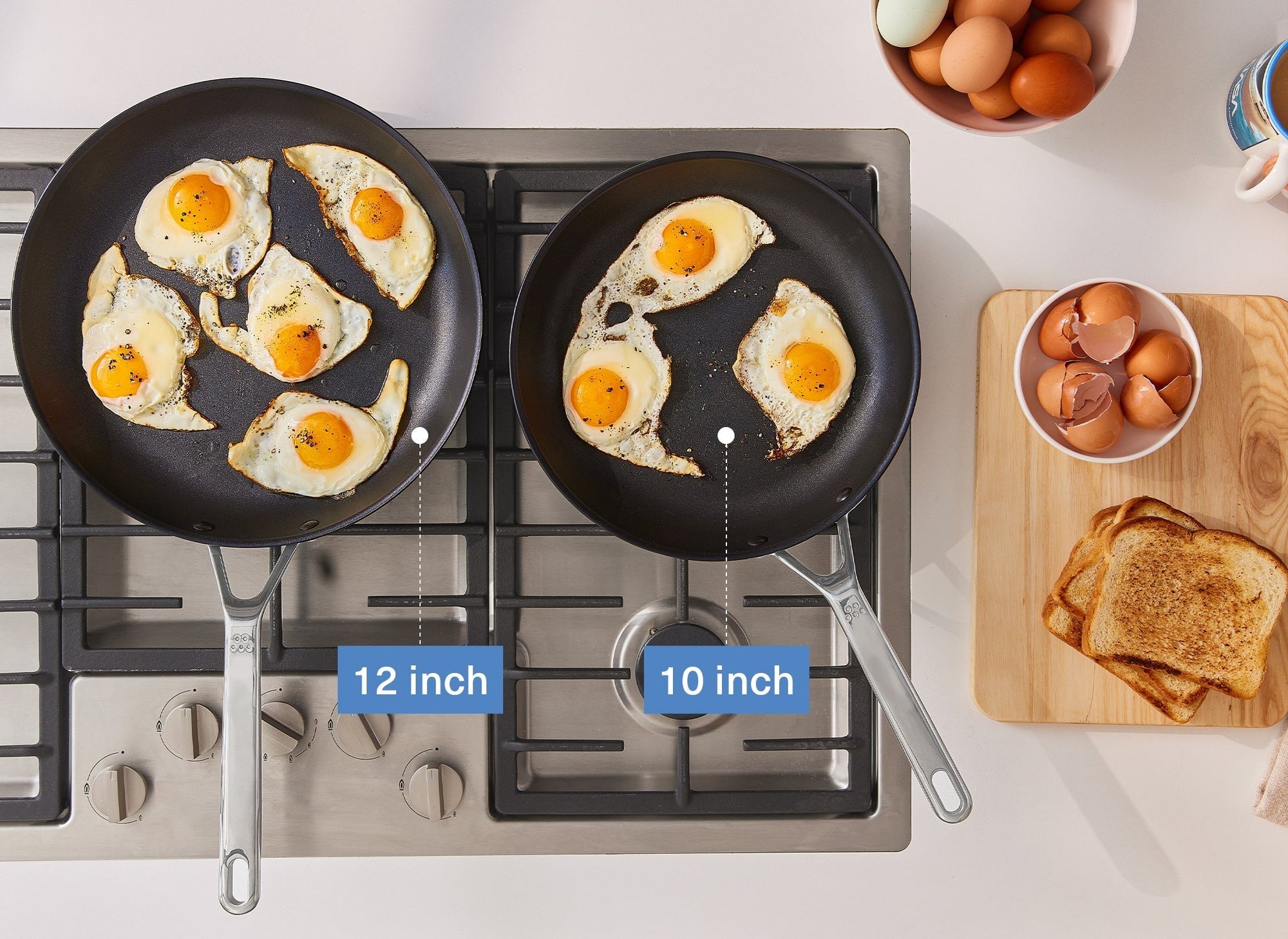 A bird’s eye view of the Misen 12-inch and 10-inch Nonstick Pans on a stovetop with fried eggs inside. To the right is a Misen Wood Cutting Board with toast and empty eggshells on it. A caption shows the 12-inch pan on the left, and the 10-inch pan on the right.