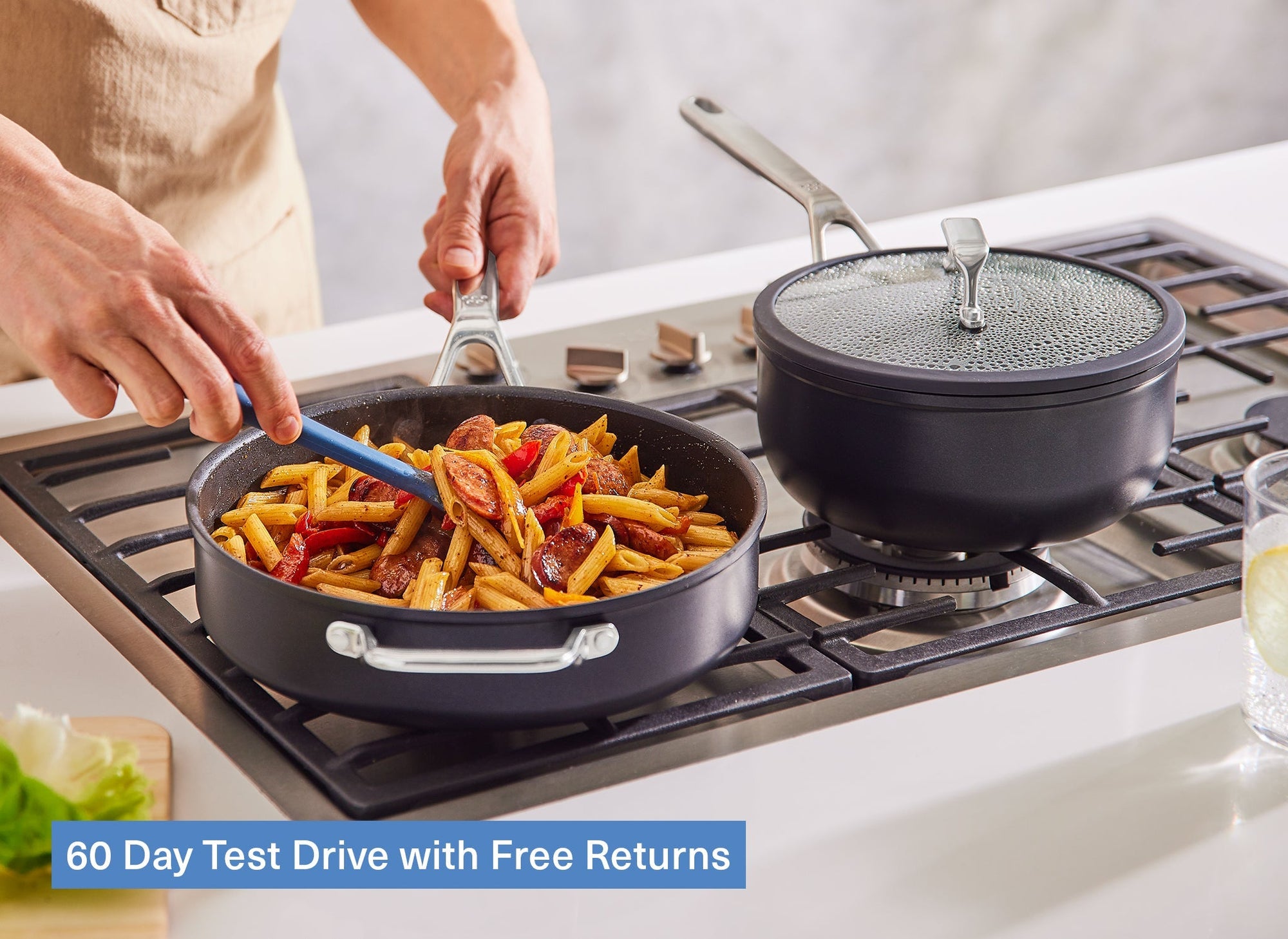 A chef’s hand uses a Misen Silicone Spoontula to stir pasta in a Misen Nonstick Sauté on a stovetop, next to a Misen Nonstick Saucier with a lid on. A caption reads “60 Day Test Drive with Free Returns.”