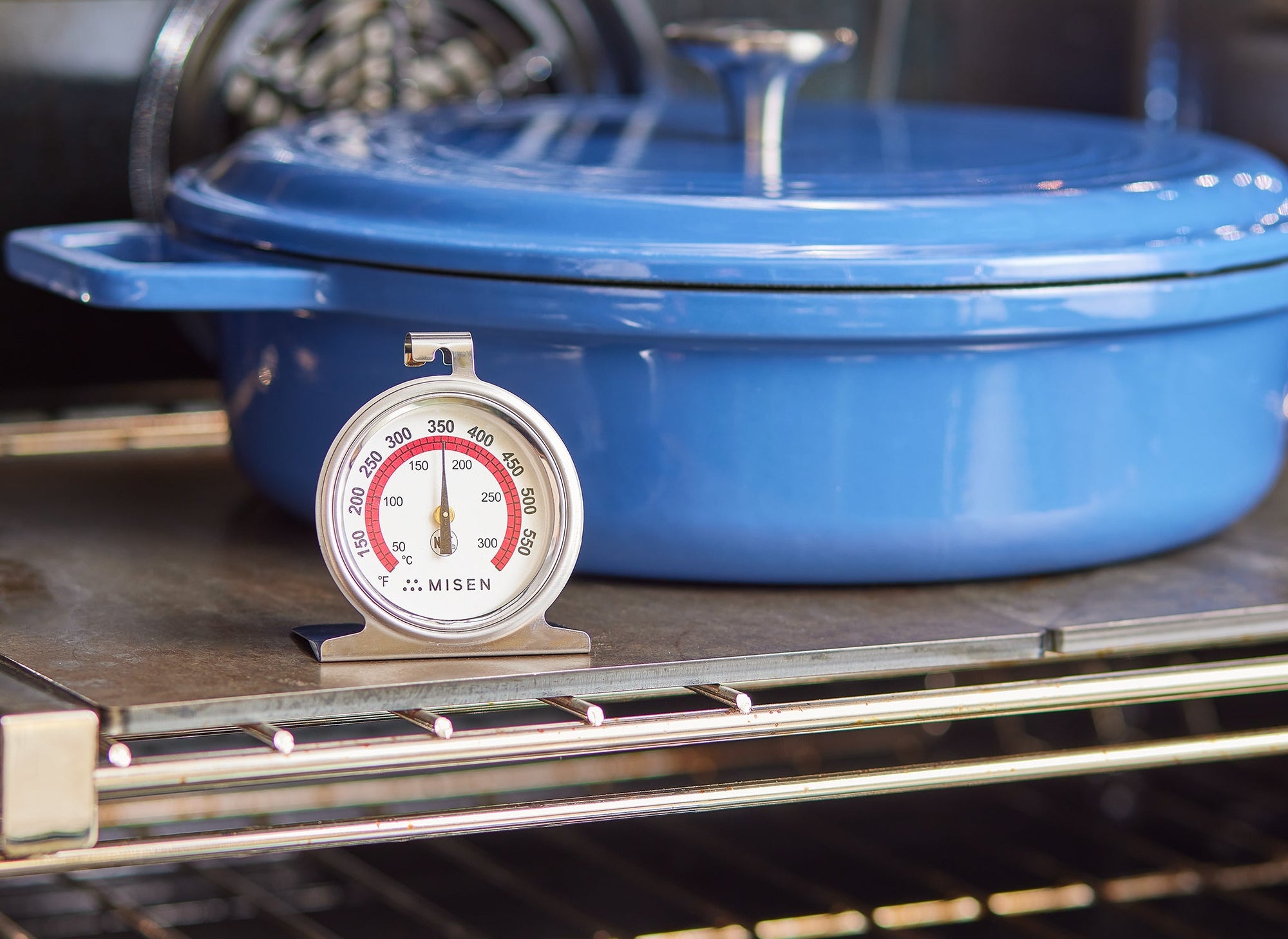 {{dial}} A stainless steel Misen Oven thermometer stands on an oven rack in front of a blue Dutch Oven. The dial reads 350 degrees Fahrenheit. 