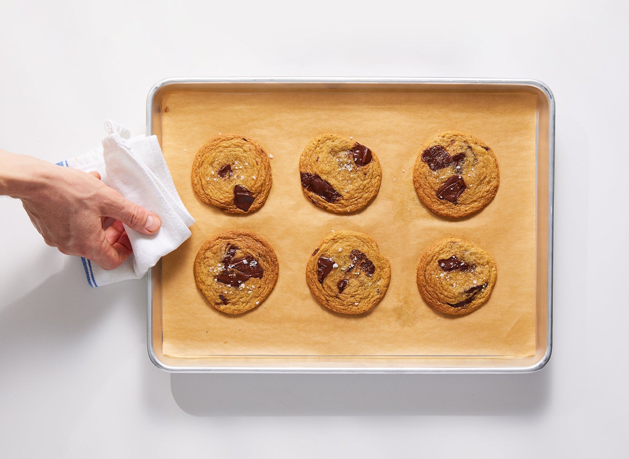 {{half-sheet,quarter-sheet,small-set,large-set}} A bird’s eye view of a Misen Sheet Pan that contains one sheet of Misen Parchment Paper beneath six baked chocolate chip cookies. A chef’s hand uses a dish towel to hold the sheet. 