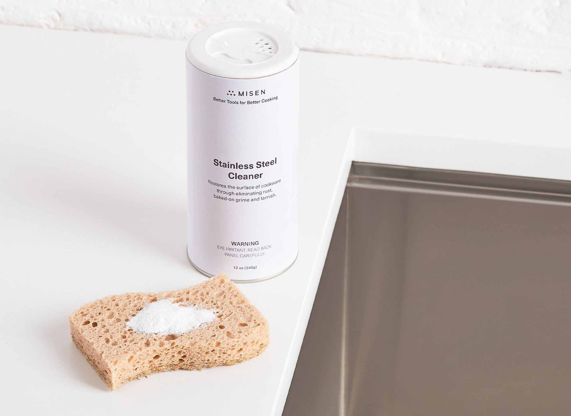 {{12-oz}} Misen Stainless Steel Cleaner canister and a sponge next to a kitchen sink, with some powder cleaner visible on the sponge surface.
