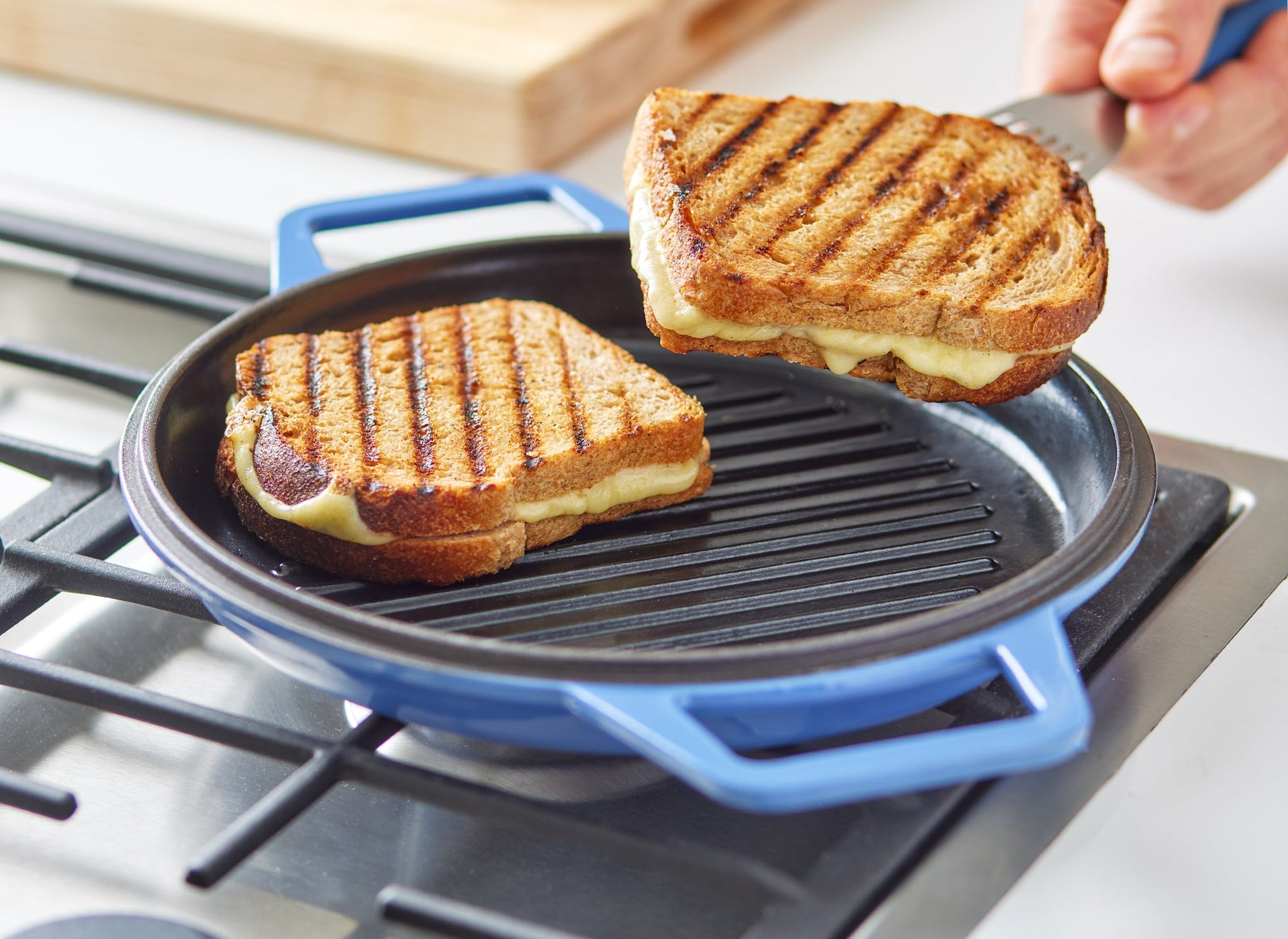 {{blue,black,gray,red,green,7-qt-blue,9-qt-blue,7-qt-black,9-qt-black,7-qt-gray,9-qt-gray,7-qt-red,9-qt-red,7-qt-green,9-qt-green}} A chef lifts a grilled cheese sandwich with perfect grill lines visible out of a Blue Misen Enameled Cast Iron Grill Pan.