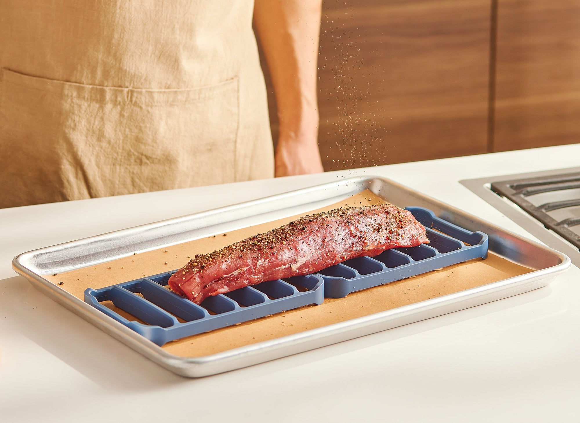 {{2-pack}} A chef seasons a pork loin, which is sitting atop two blue Misen Silicone Roasting Racks in a parchment paper-lined baking pan.