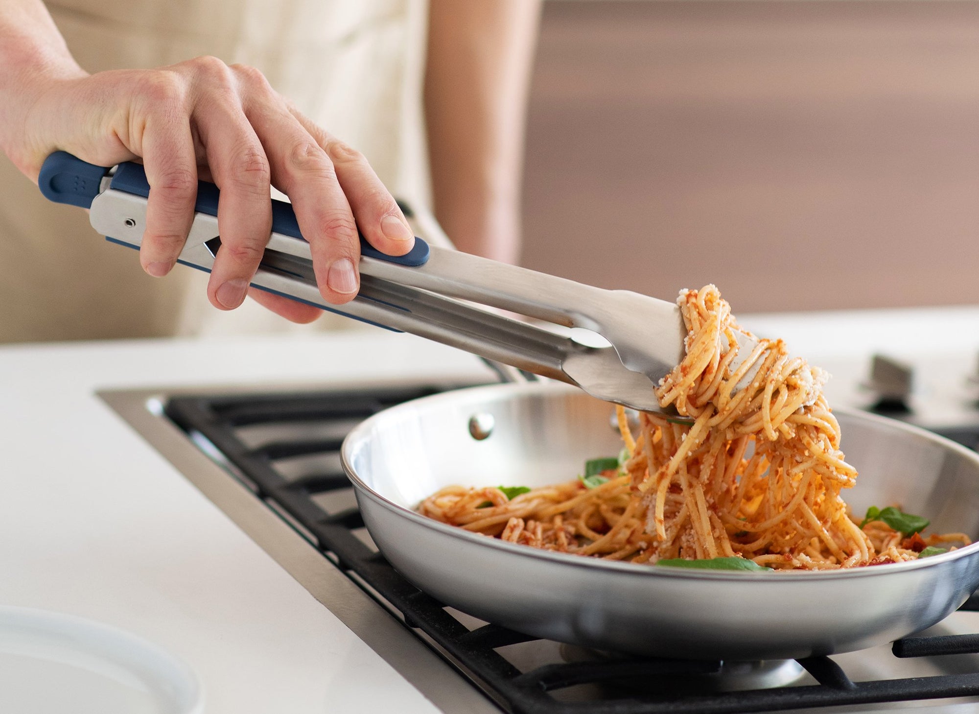 {{stainless-steel-blue, stainless-steel-black,stainless-steel-gray}} Blue Misen Metal Tongs lifting spaghetti with marinara sauce and basil out of a stainless steel skillet.
