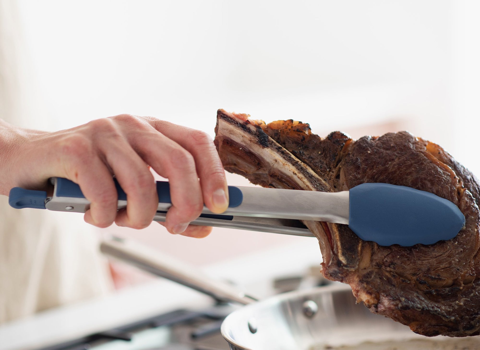 {{silicone-blue,silicone-black,silicone-gray}} Blue Misen Silicone Tongs holding a cooked ribeye steak above a stainless steel skillet.