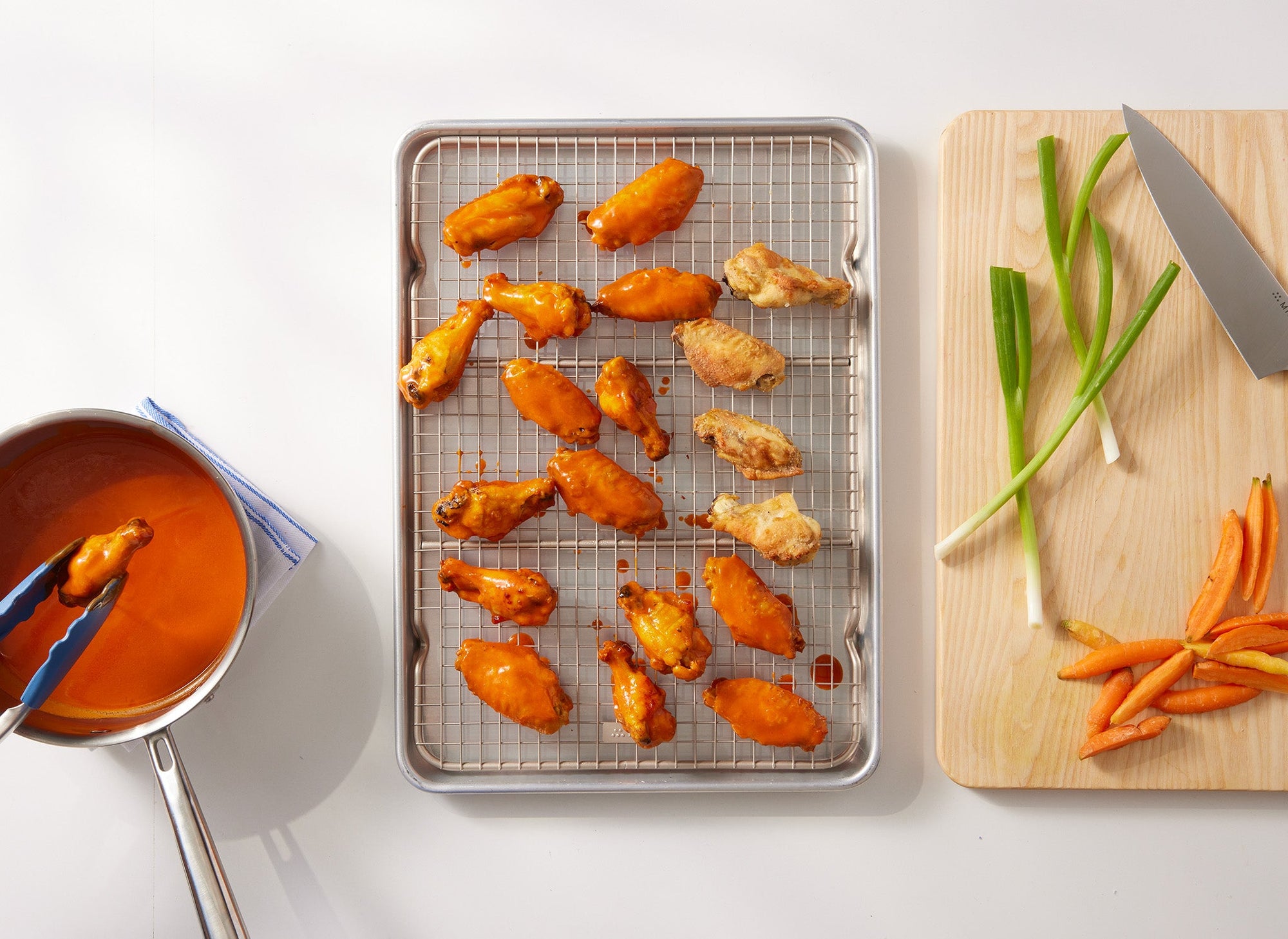 {{quarter-sheet,half-sheet,small-set,large-set}} A bird’s eye view of a Misen Wire Rack nestled within a Misen Sheet Pan, next to a Misen Wood Cutting Board. Atop the Wire Rack are cooked chicken wings that have been dunked in a visible pot of red sauce. Atop the cutting board is a Misen Chef’s Knife and assorted vegetables. 