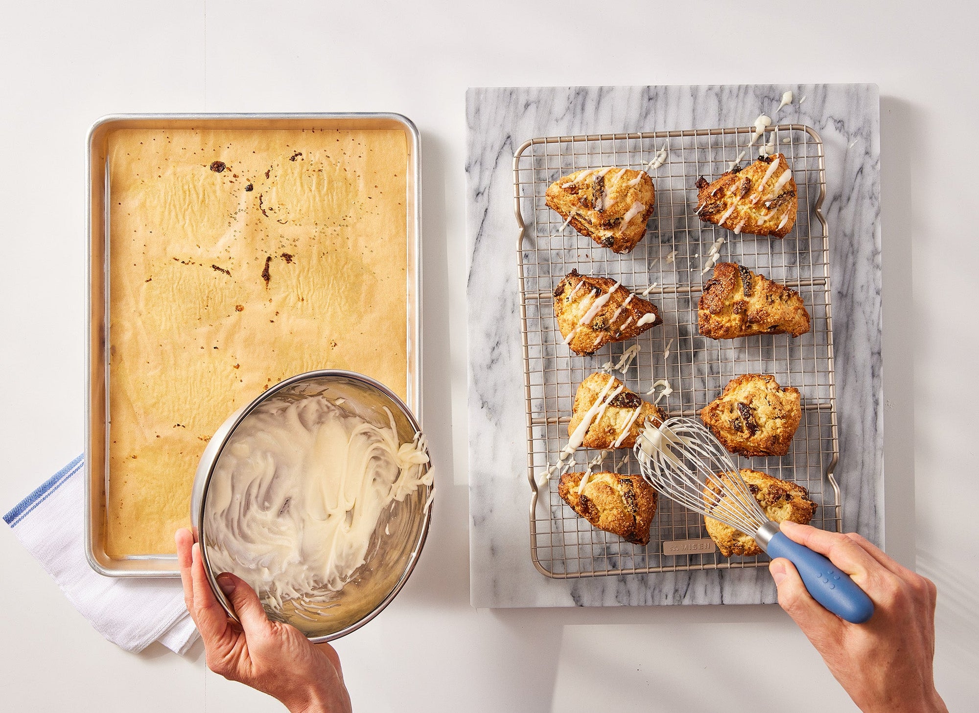 {{quarter-sheet,half-sheet,small-set,large-set}} A pair of hands uses a Misen Spatula scoop icing from a metal bowl to drizzle icing on baked scones, which sit atop a Misen Wire Rack, which is next to a Misen Sheet Pan lined with Misen Parchment Paper.
